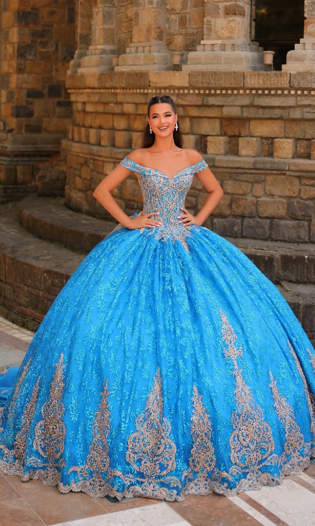 A young woman poses outdoors before a stone building, wearing the Quinceanera Dress 54324 By Amarra—an off-the-shoulder gown with gold lace embroidery and a voluminous blue skirt.